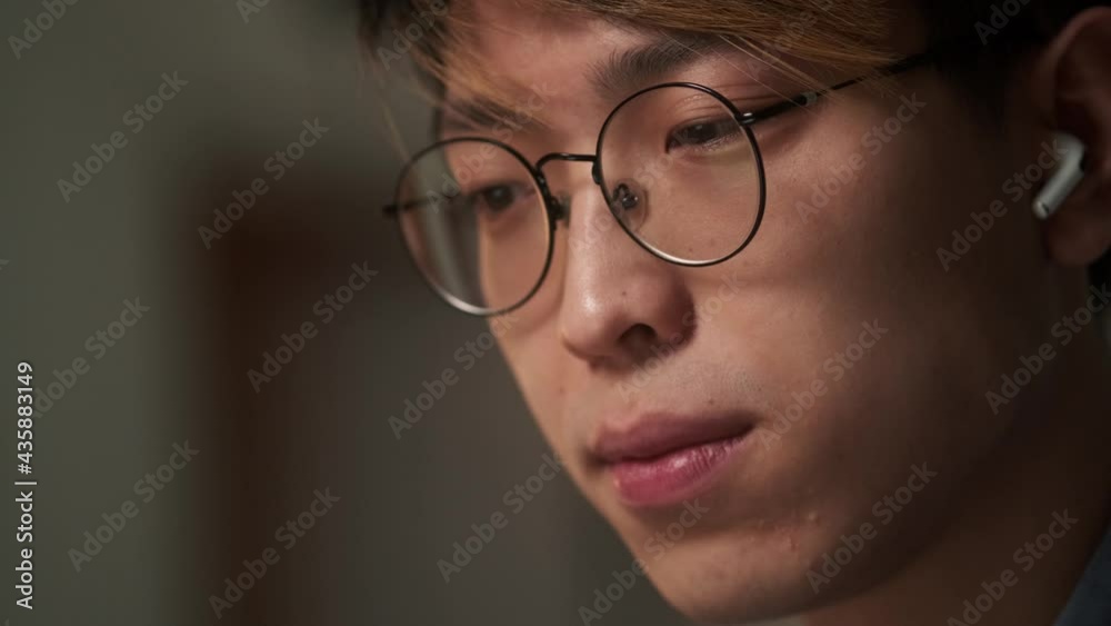 The close-up view of the chinese man in the earbuds shaking his head positively while sitting in the night office The close-up view of the chinese man in the earbuds shaking his head positively while sitting in the night office