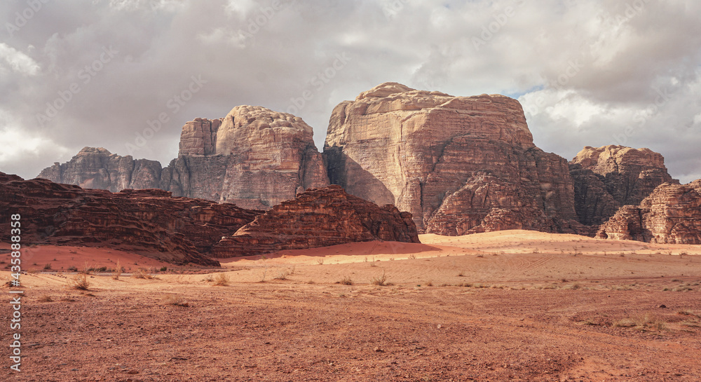 Rocky massifs on red sand desert, few dry grass cluster ground, cloudy sky in background, typical scenery in Wadi Rum, Jordan Rocky massifs on red sand desert, few dry grass cluster ground, cloudy sky in background, typical scenery in Wadi Rum, Jordan