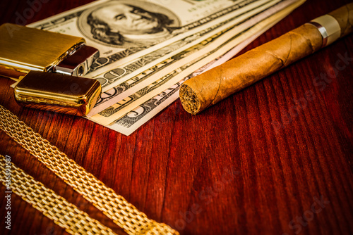 Cuban cigar and a gold chain with money and golden lighter on a table in mahogany. Focus on the cuban cigar