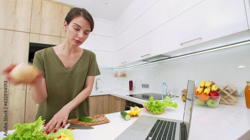 Sliding view of attractive brunette woman cutting vegetables, putting them on the plate and having webcam chat to someone using a laptop