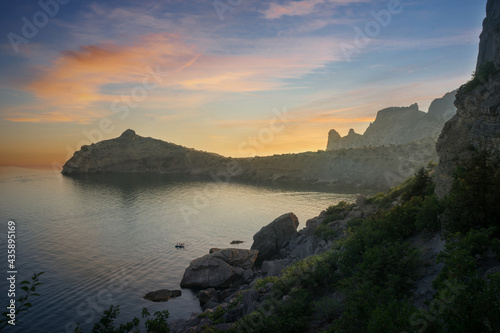 Sunset over a beautiful cape on the Black Sea in Crimea in Russia. Cape Kapchik in the evening before sunset. High cliffs surround Blue Bay. A beautiful bay and people boating. Quiet calm weather