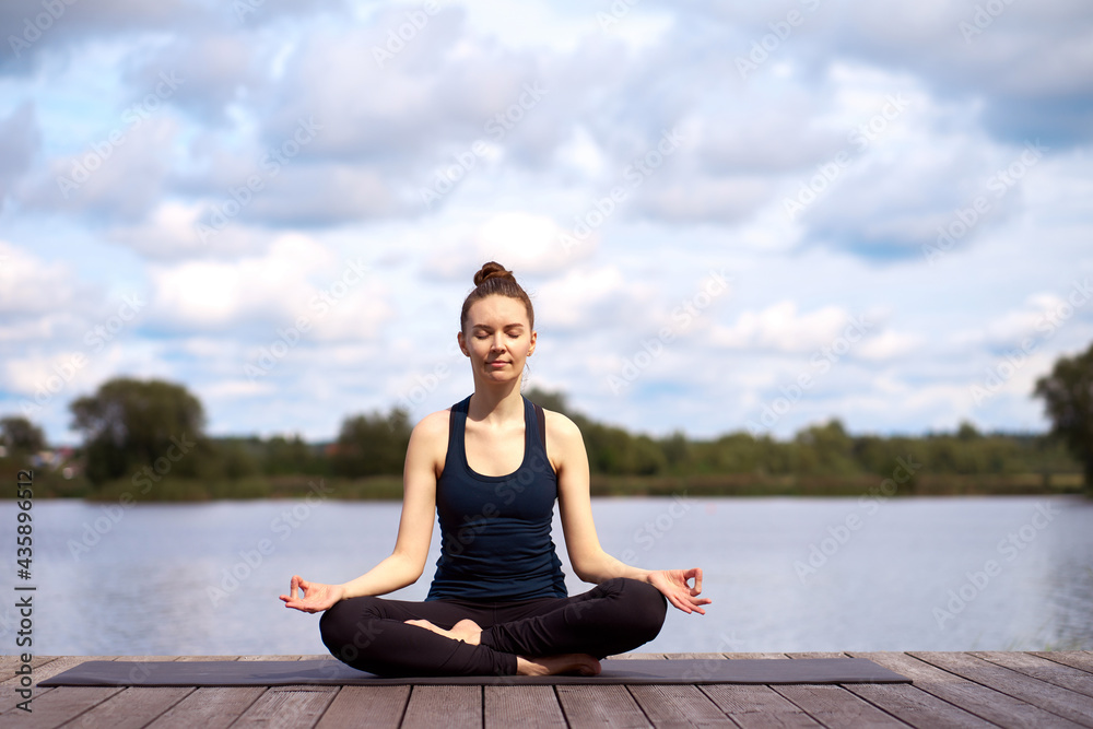 Beautiful young woman with closed eyes relaxing and practicing yoga on the wooden pier near lake. Outdoor activities.