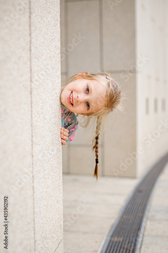A blonde girl with a pigtail sticks her head out from behind a pillar
