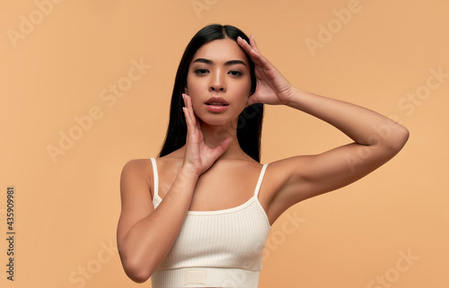 Young Asian woman with clean healthy glowing skin in white top isolated on beige background. Facial skin care concept, spa, cosmetology, plastic surgery.