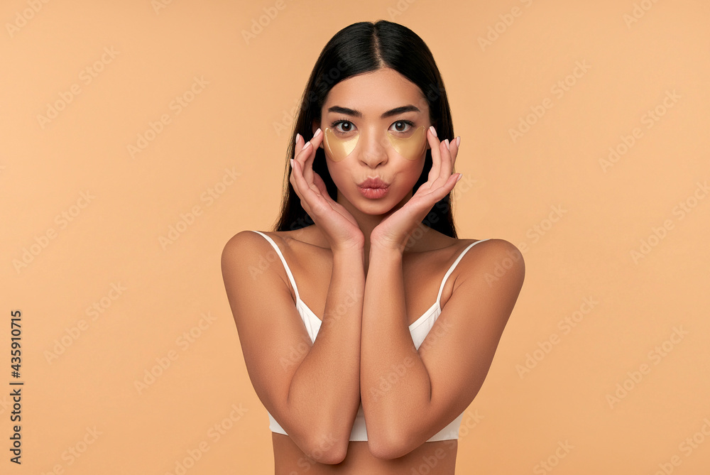 Fototapeta premium Young Asian woman in white lingerie and clean radiant skin with moisturizing patches under the eyes on a beige background. Spa care, cosmetology.