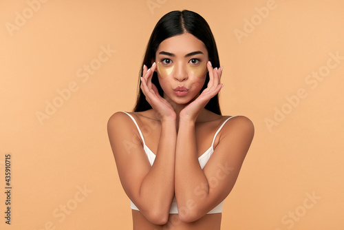 Young Asian woman in white lingerie and clean radiant skin with moisturizing patches under the eyes on a beige background. Spa care, cosmetology.