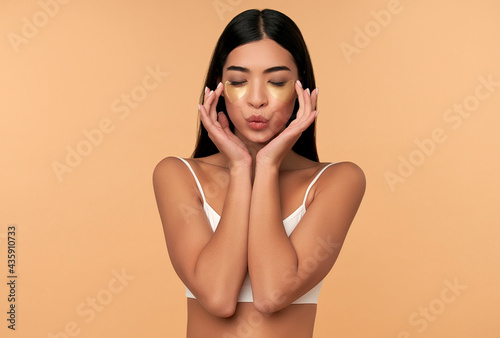Foto Young Asian woman in white lingerie and clean radiant skin with moisturizing patches under the eyes on a beige background