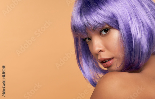 Young Asian woman with clean radiant skin in a purple wig and bright hair on a beige background. Spa care, facial skin care, beauty cosmetology.