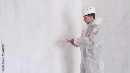 A man in a protective suit and helmet holds a spatula in his hand. Leveling the walls with a spatula and plaster. Updates in the house. The concept of home renovation.