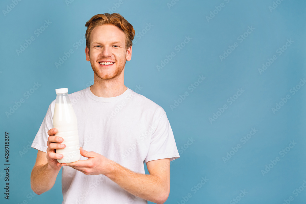 Smiling cheerful handsome young bearded man 20s wearing casual clothes posing holding in hand vegan milk isolated over blue color wall background. Smiling cheerful handsome young bearded man 20s wearing casual clothes posing holding in hand vegan milk isolated over blue color wall background.