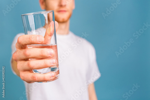 A young redhead man in a white t-shirt isolated over blue background drinking water from a glass.