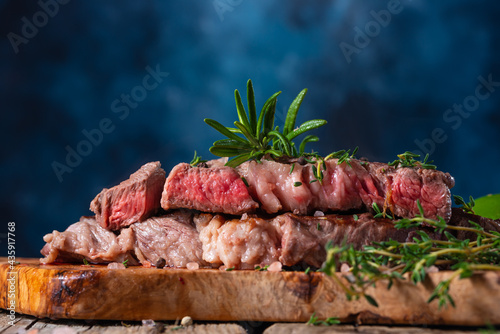 In the photo we see a slice of bread with steak. Top of the steak is decorated with sprigs of fresh herbs. In the background is a photo of a dark blue smoky background. Close-up.