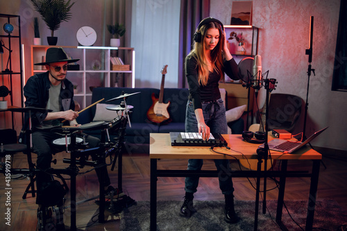 Stylish man in sunglasses and hat playing on electronic drums while charming woman in headphones performing on synthesizer. Group of two musicians recording music in studio with dark atmosphere.