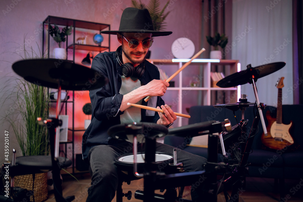 Fototapeta premium Happy stylish man sitting at dark music studio and playing electronic drums with sticks. Professional musician wearing trendy black hat and sunglasses.