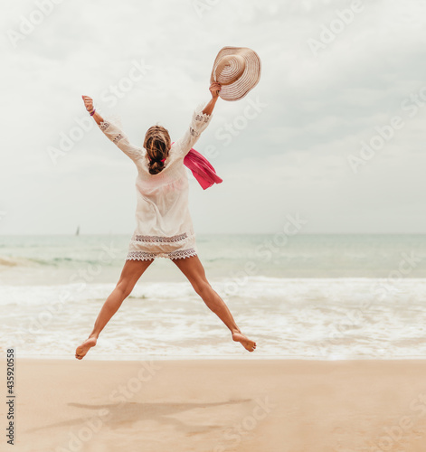 woman jumping on the shore of the beach with hat in hand. she stays slim and jumps with ease. she is happy celebrating something. post pandemic vacation. vaccine. © Karlos Garciapons