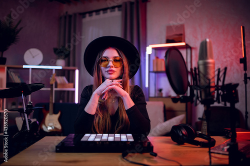 Portrait of stylish young woman in trendy black hat and sunglasses looking at camera while sitting at table with various music instruments. Female musician at home studio.