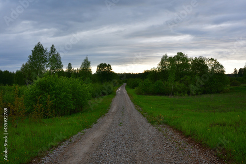 After the rain, a gravelled field road descends past green meadow bushes and into the forest on the horizon, beyond which the reflections of the setting sun illuminate the cloudy evening sky.