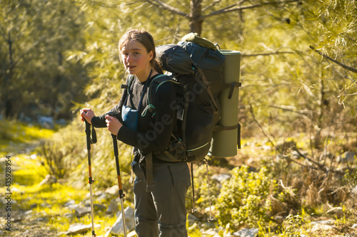 A beautiful smiling girl in the rays of the sun holds trekking poles and walks along a mountain trail. A young girl in thermal underwear and trekking pants with a backpack goes hiking in the forest.