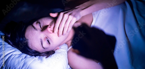 A young girl lies in bed and cannot sleep, reads and chat on social networks on a smartphone. A woman with insomnia is watching a video online on a device.