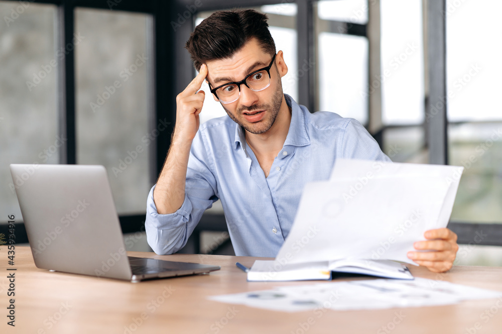 Shocked frustrated young caucasian business leader, top manager or freelancer wearing glasses sits at the workplace in the office, looks with surprise at business documents, puzzled Shocked frustrated young caucasian business leader, top manager or freelancer wearing glasses sits at the workplace in the office, looks with surprise at business documents, puzzled
