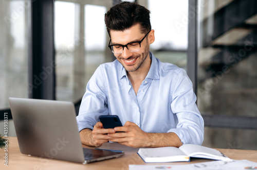 Joyful caucasian young man, manager or freelancer sitting at his desk in the office, texting with a friend or client using a smartphone, online chat, browsing the Internet, answering email, smiling