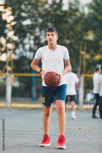 Cute boy in white t shirt plays basketball on a city playground. Active teen enjoying outdoor game with orange ball. Hobby, active lifestyle, sport for kids. 