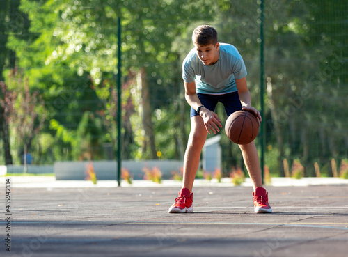 Cute boy in green t shirt plays basketball on a city playground. Active teen enjoying outdoor game with orange ball. Hobby, active lifestyle, sport for kids.  © Natali