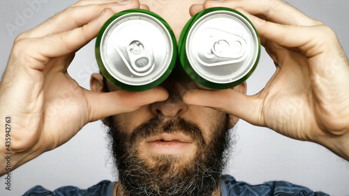 A young bearded man holds two cans of beer to his eyes pretending to look through binoculars