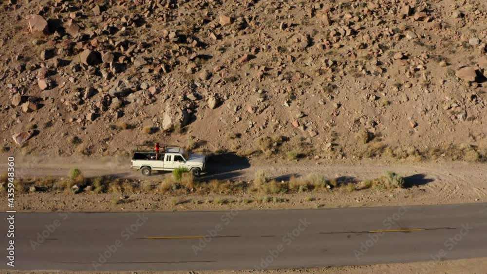 Pickup driving un gravel road in desert landscape with person standing on the back bed. Panning drone view.