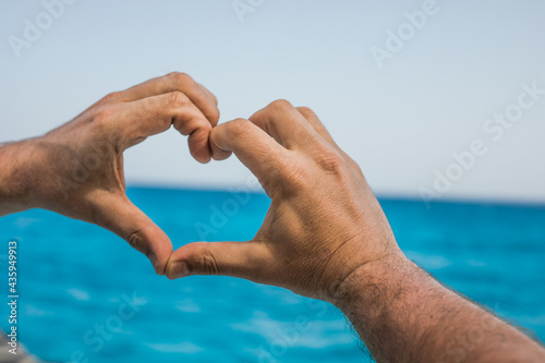 The hands of a man  depict a sign or symbol of heart and love against the backdrop of the sea and the sky. © Maria