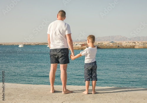 Father and son spending time together sea vacation. Little boy and Dad stay on beach and looks out to sea. Back view. Family with one child. Happy childhood with daddy.