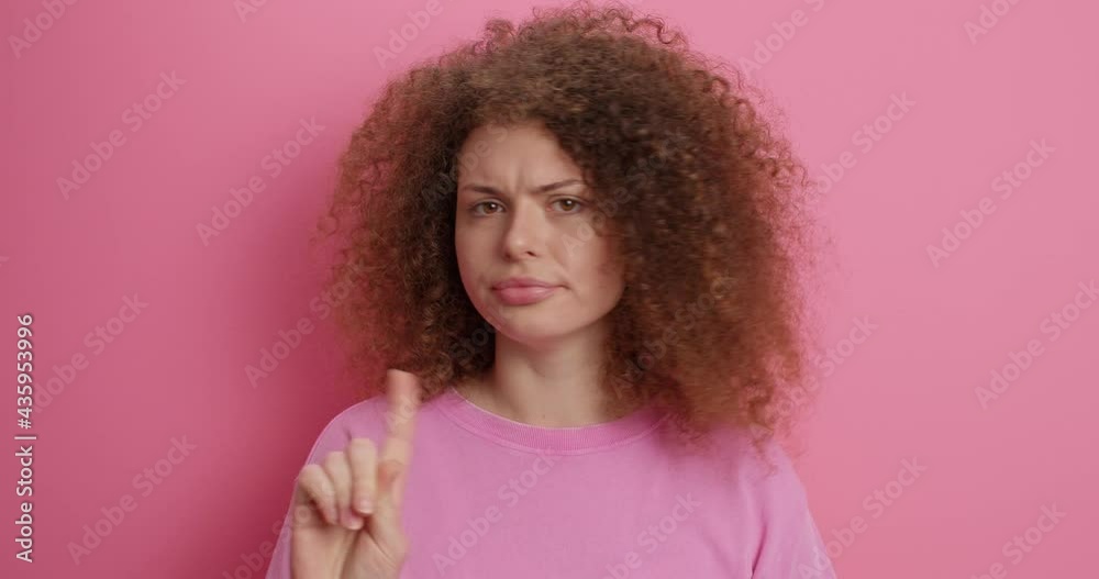 Serious curly haired woman disagrees with you shakes index finger says no dressed casually has displeased expression dressed in casual clothes isolated over pink background doesnt allow something