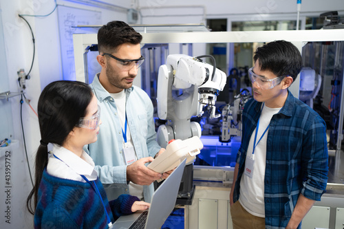 Caucasian male engineer standing hold a robot controller next to Asian female apprentice holding a laptop, listen intently to young Asian male CEO after demonstrated the work of robotic machine to him