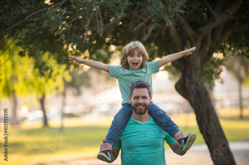 Portrait of a young dad and his child in the park. Happy kid son stretching out hands while his father carrying him on shoulders.