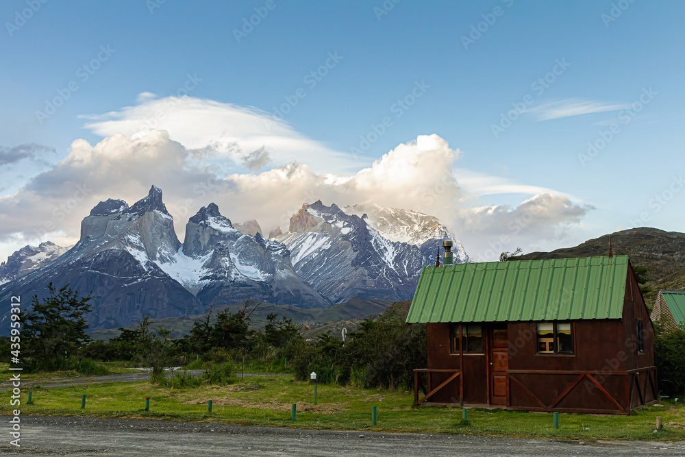 Pequena casa de madeira com gramado na frente e ao fundo a bela vista da cordilheira do parque nacional Torres del Paine ao entardecer