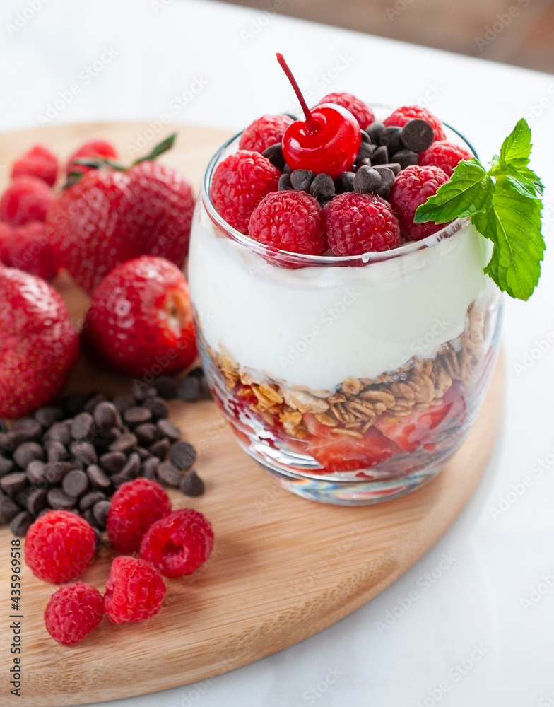 A glass of yogurt with raspberries granola, strawberries and chocolate chips with a mint or peppermint leaf on top over a wooden cutting board in a white background A glass of yogurt with raspberries granola, strawberries and chocolate chips with a mint or peppermint leaf on top over a wooden cutting board in a white background