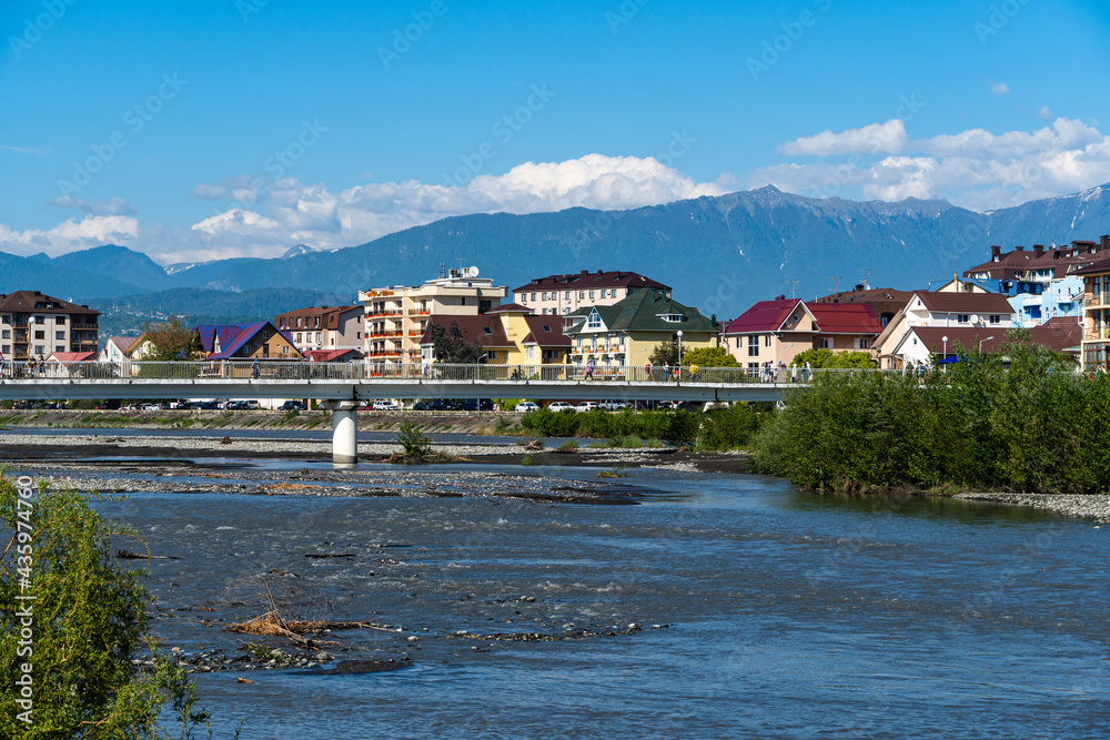 Naklejka premium Residential buildings in federal territory of Sirius against backdrop of snow-capped peaks. Caucasus mountains. Imeretinskaya lowland. In foreground is Mzymta.Adler. Sochi, Russia - May 18, 2021