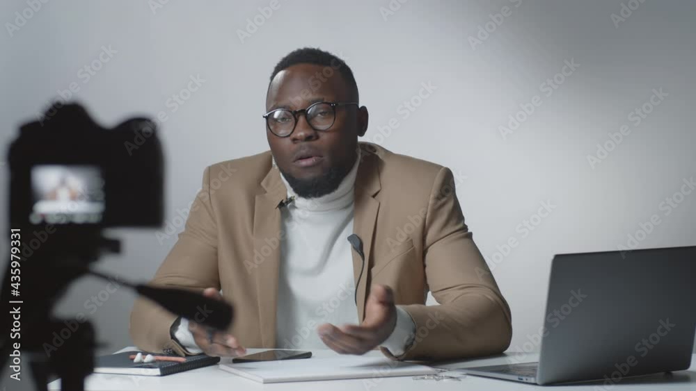 Selective focus shot young Afro-American man sitting at desk with laptop and explaining something on camera while filming vlog in studio Selective focus shot young Afro-American man sitting at desk with laptop and explaining something on camera while filming vlog in studio