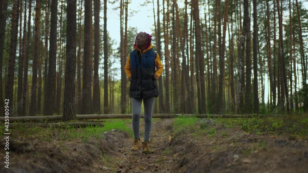 Beautiful black girl in yellow jacket with blue backpack got lost walking in woods on sunny day. Lonely woman looks around in forest. Beautiful black girl in yellow jacket with blue backpack got lost walking in woods on sunny day. Lonely woman looks around in forest.