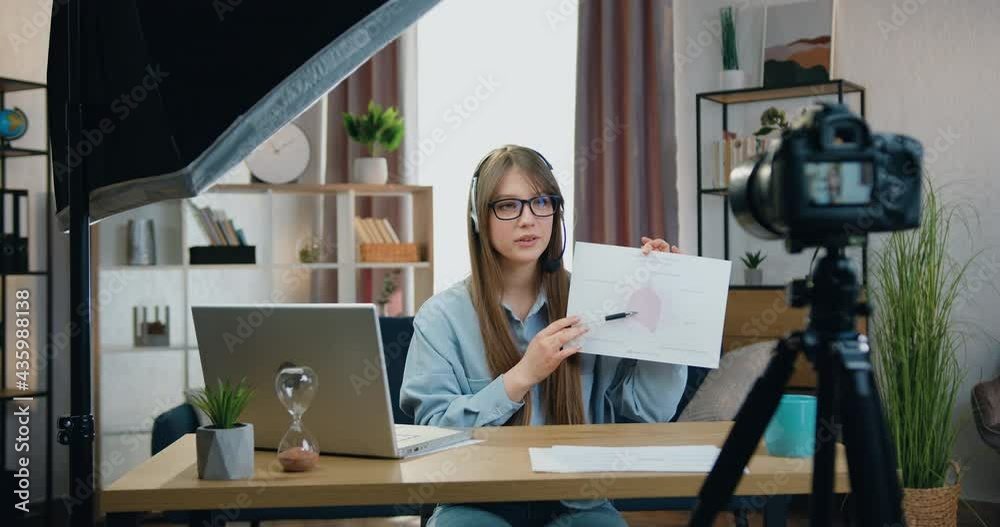 Remote learning concept where attractive confident skilled young girl in headphones sitting in front of camera in home office and holding online conference explaining report with charts