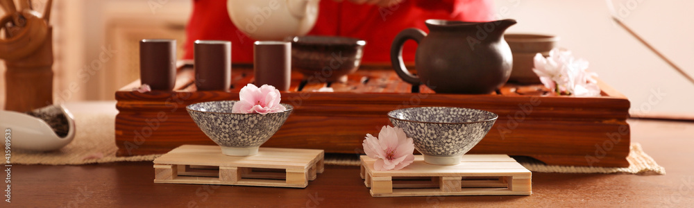Closeup view of master conducting traditional ceremony at table indoors, focus on cups and sakura flowers. Banner design