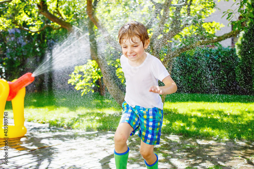 Funny little kid boy playing with a garden hose sprinkler on hot and sunny summer day. Child having fun with sprinkling water, drops rain. Outdoors leisure wth water for children. Rain rubber boots.