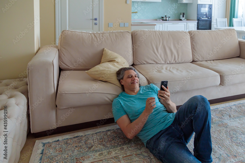 An adult man browses social networks on a mobile phone while sitting on the floor in an apartment. An adult man browses social networks on a mobile phone while sitting on the floor in an apartment.