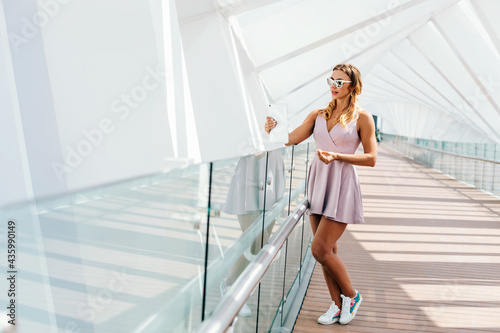 A young woman wearing sunglasses and a summer dress smiles happily while making a video call with a tablet while walking