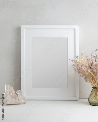 Empty white frame, glass vase with dried flowers and natural crystal in a light interior. Mock up poster with place for your design.