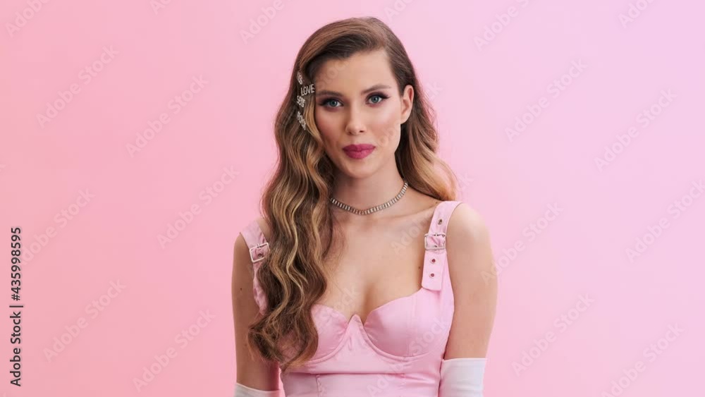 A beautiful young woman wearing gorgeous evening pink dress with gloves and hairpins is posing to the camera standing isolated over a pink wall in the studio A beautiful young woman wearing gorgeous evening pink dress with gloves and hairpins is posing to the camera standing isolated over a pink wall in the studio