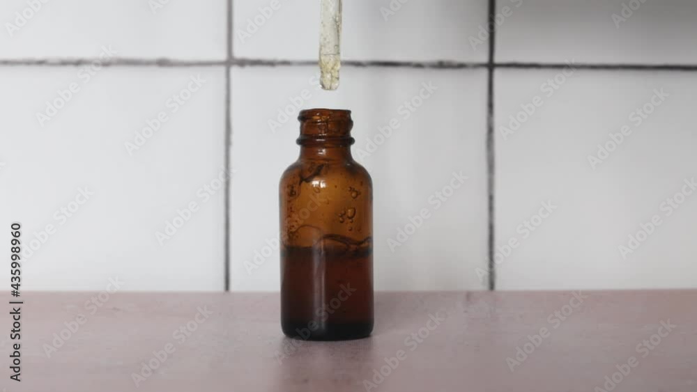 Pipette with yellow natural oil dropping over glass bottle on a white bathroom tile background. Pipette with yellow natural oil dropping over glass bottle on a white bathroom tile background.