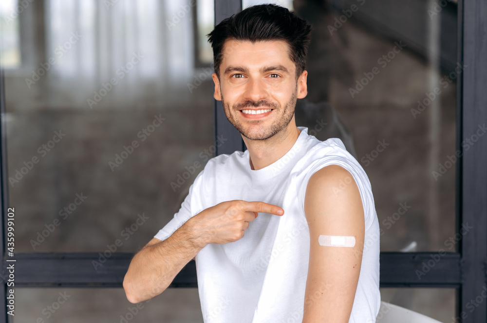 Obraz premium Happy healthy young Caucasian male, in casual clothes, received a vaccine against covid 19, immunity from viruses and infections, take care of his health, points to a band-aid on his shoulder, smiling