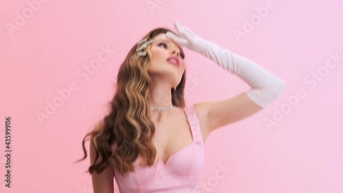 A beautiful young woman wearing gorgeous evening pink dress with gloves and hairpins is losing consciousness isolated over a pink wall in the studio