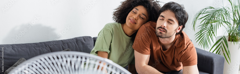 Fototapeta premium pleased interracial couple sitting on couch near blurred electric fan in living room, banner